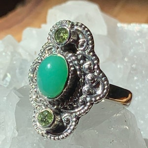 May include: A silver ring with an oval green gemstone and two smaller green gemstones. The ring has a detailed filigree design.