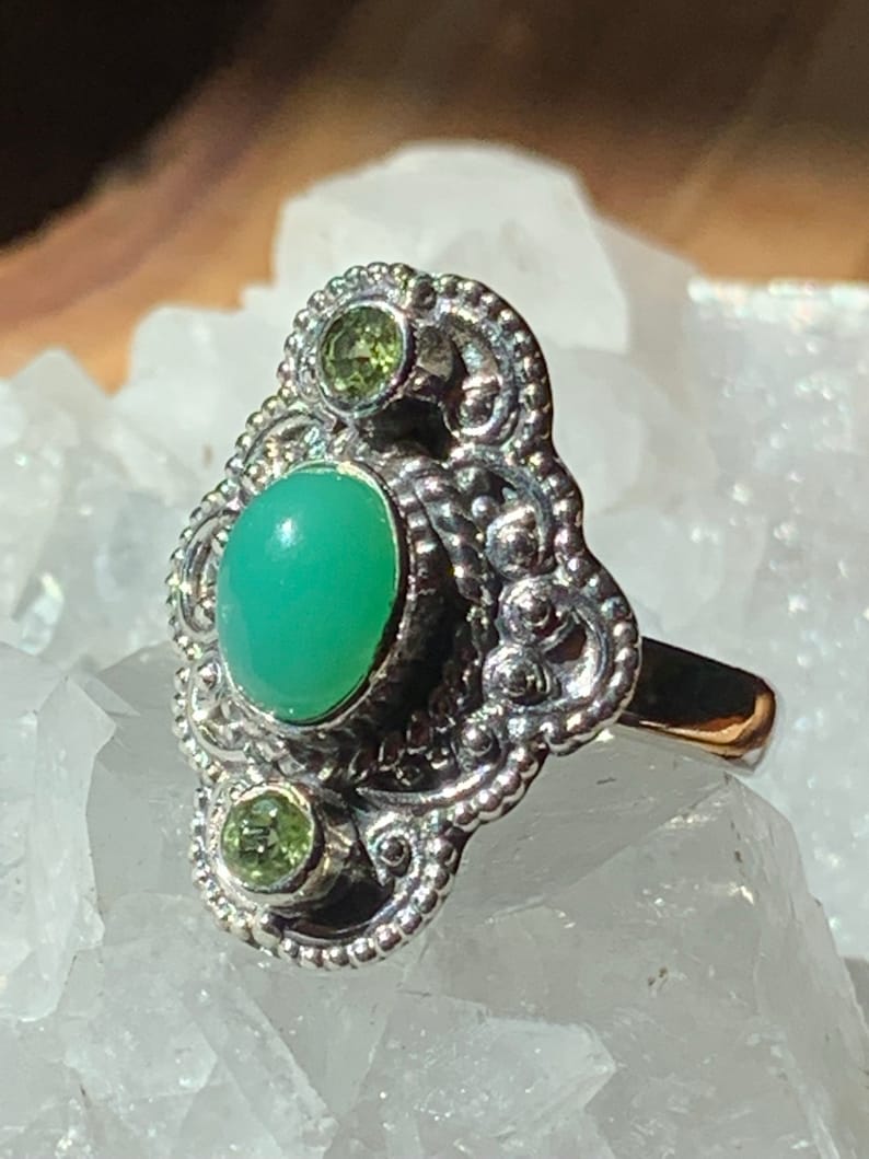 May include: A silver ring with an oval green gemstone and two smaller green gemstones. The ring has a detailed filigree design.