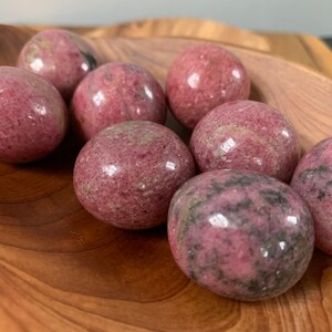 May include: A collection of eight pink and gray tumbled rhodonite spheres in a wooden bowl. The spheres are smooth and polished, with a variety of patterns and textures.