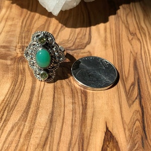 May include: A silver ring with a green oval gemstone surrounded by smaller green gemstones. The ring is on a wooden surface next to a US quarter.