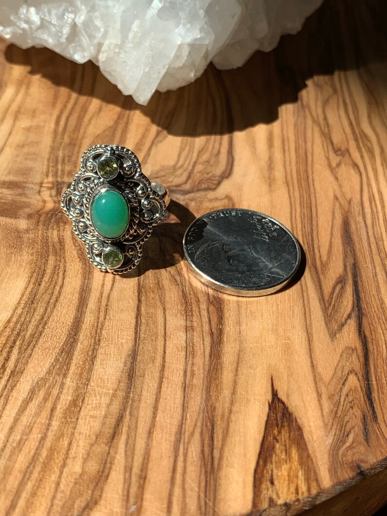 May include: A silver ring with a green oval gemstone surrounded by smaller green gemstones. The ring is on a wooden surface next to a US quarter.