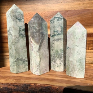 May include: Four green and white polished crystal points, also known as wands, are arranged on a wooden surface. The points are all different sizes and shapes, but they are all made of the same type of crystal.