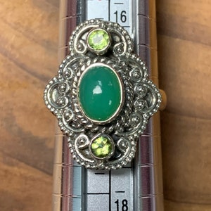 May include: A silver ring with a green oval gemstone and two smaller green gemstones. The ring is set on a silver band with intricate details. The ring is sized on a ring sizer that shows size 16.