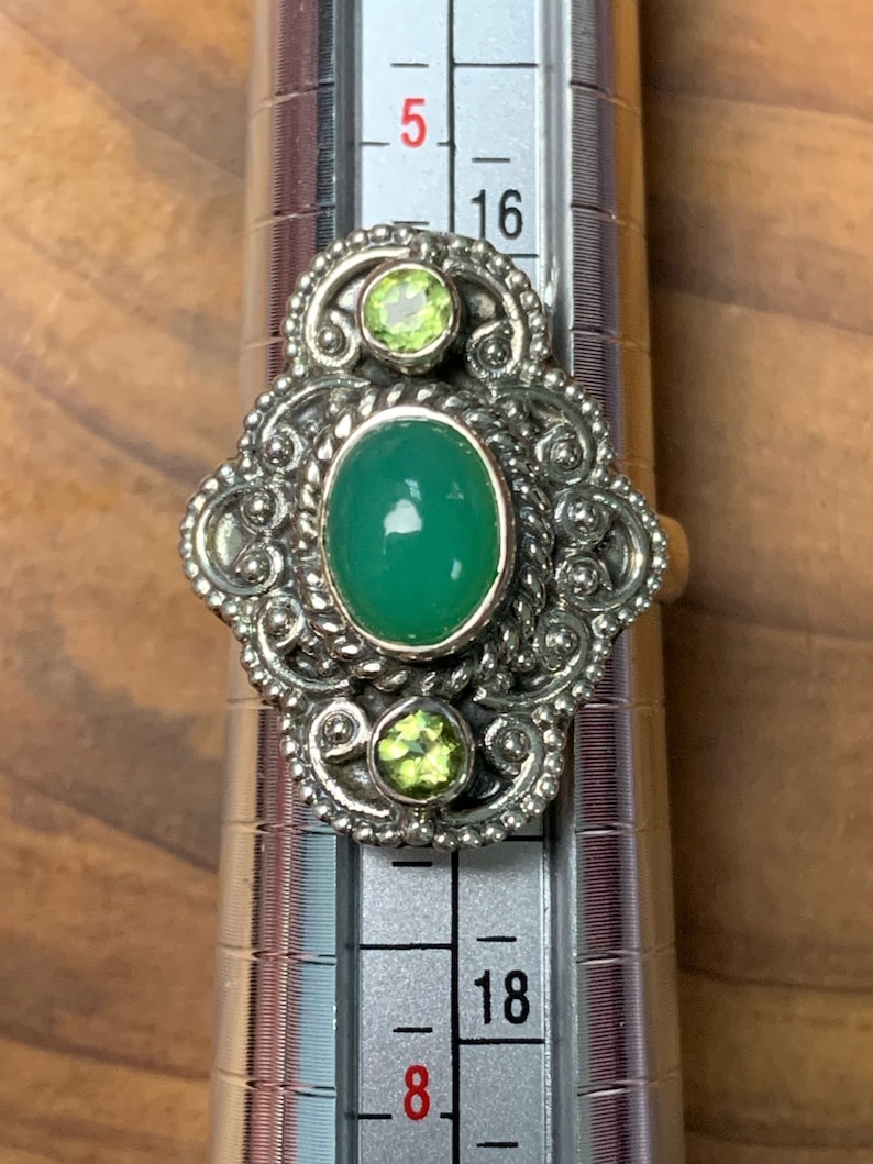 May include: A silver ring with a green oval gemstone and two smaller green gemstones. The ring is set on a silver band with intricate details. The ring is sized on a ring sizer that shows size 16.