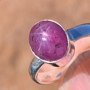 May include: A silver ring with a pink star ruby gemstone. The ruby is oval shaped and has a star-like pattern.
