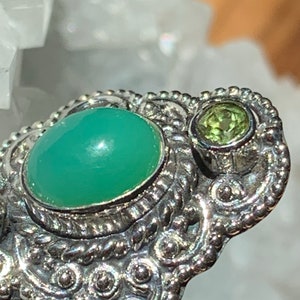 May include: A silver ring with a green gemstone and a smaller green gemstone. The ring has a detailed, ornate design.