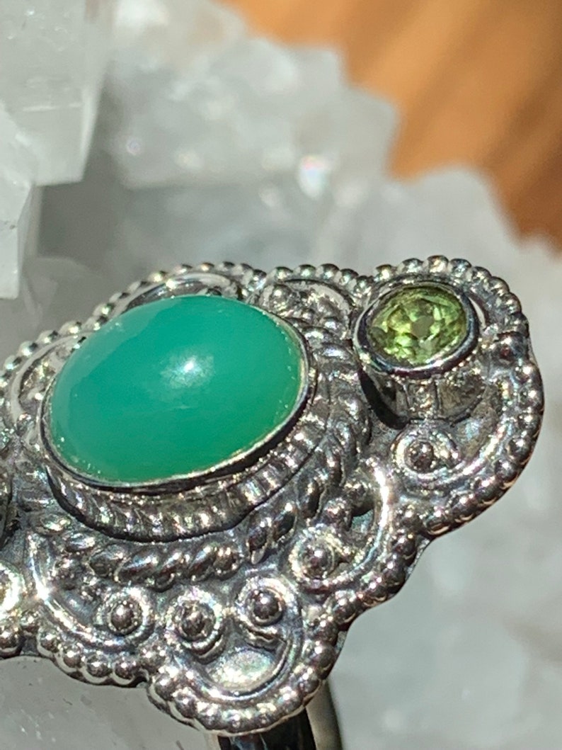 May include: A silver ring with a green gemstone and a smaller green gemstone. The ring has a detailed, ornate design.