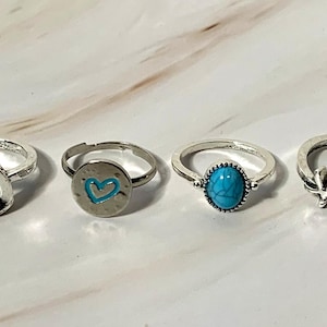 May include: Six silver rings with various designs are displayed. The rings include a chevron-style ring, a teardrop-shaped ring, a ring with a blue heart, a ring with a turquoise stone, a starfish ring, and a ring with a chain-link design.