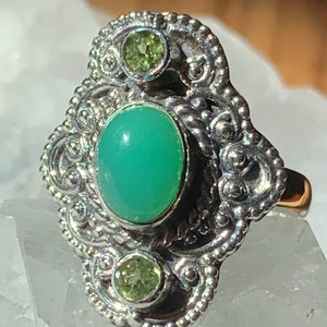 May include: A silver ring with a green gemstone and two smaller green gemstones. The ring has a detailed design with intricate swirls and beading.