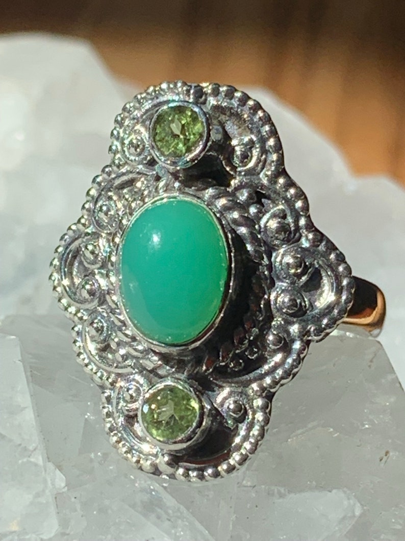 May include: A silver ring with a green gemstone and two smaller green gemstones. The ring has a detailed design with intricate swirls and beading.