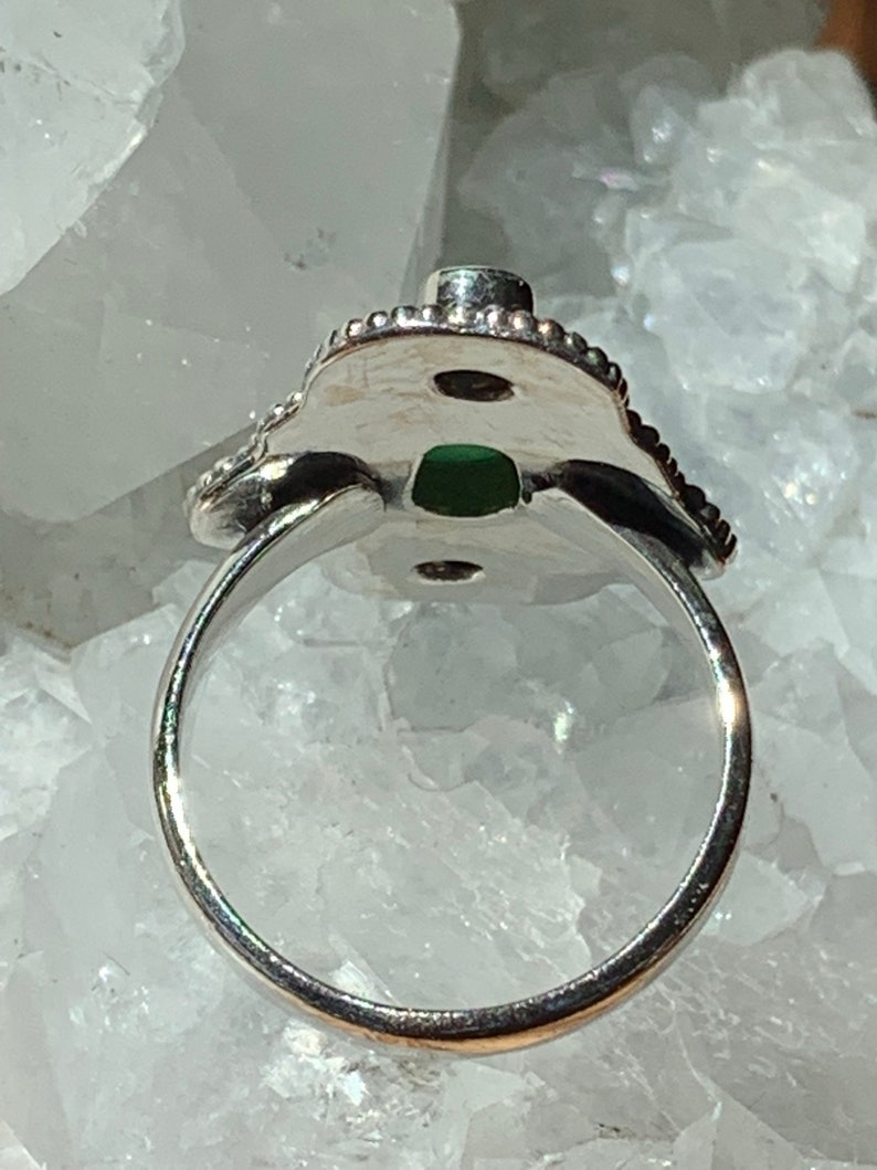 May include: A silver ring with a green stone set in a decorative, ornate design. The ring is open on the back, allowing for easy adjustment.
