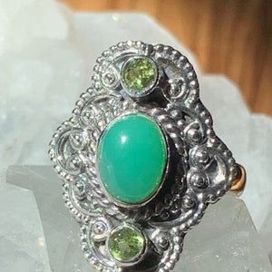 May include: A silver ring with a green oval gemstone and two smaller green gemstones. The ring has a detailed, ornate design.