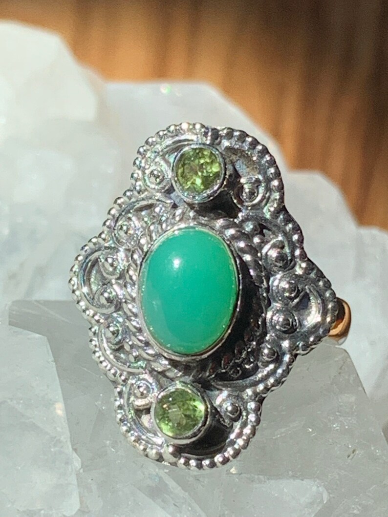 May include: A silver ring with a green oval gemstone and two smaller green gemstones. The ring has a detailed, ornate design.