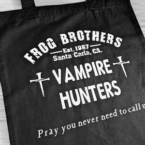 May include: Black tote bag with white text. The text reads "FROG BROTHERS - Est. 1987 Santa Carla, CA. VAMPIRE HUNTERS" with cross symbols and the phrase "Pray you never need to call us."