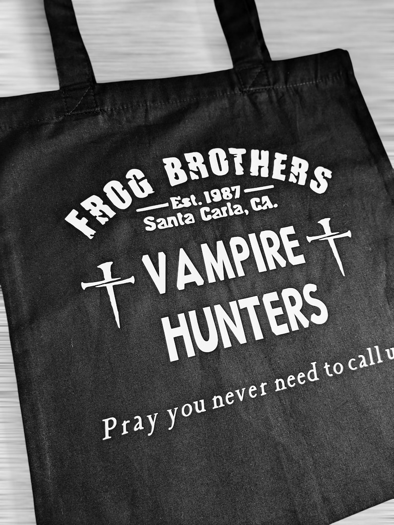 May include: Black tote bag with white text. The text reads "FROG BROTHERS - Est. 1987 Santa Carla, CA. VAMPIRE HUNTERS" with cross symbols and the phrase "Pray you never need to call us."