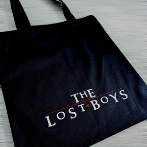 May include: Black tote bag with the text "THE LOST BOYS" in white, with a red line and dot. The bag has black handles and is made of a sturdy fabric. The design is centered on the front of the bag.