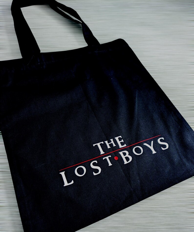 May include: Black tote bag with the text "THE LOST BOYS" in white, with a red line and dot. The bag has black handles and is made of a sturdy fabric. The design is centered on the front of the bag.