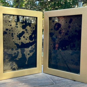 Pode incluir: Uma moldura dourada com dobradiças e dois painéis. Cada painel apresenta um fundo escuro com salpicos dourados. A moldura está aberta, revelando a obra de arte. A moldura é feita de metal e tem um design simples e moderno.