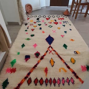 Puede incluir: Una alfombra marroquí blanca con patrones geométricos coloridos y flecos. La alfombra tiene una forma de diamante en el centro con un diamante azul en el medio.