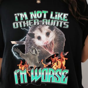 May include: A black t-shirt featuring a graphic print of a screaming opossum and a small raccoon surrounded by orange flames and blue lightning. The text reads I'M NOT LIKE OTHER AUNTS I'M WORSE in a bold, light blue, outlined font style.