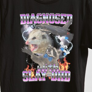 May include: Black t-shirt featuring a graphic print of a possum and a raccoon. The text "DIAGNOSED with SLAY-DHD" is printed in pink and purple on the shirt.