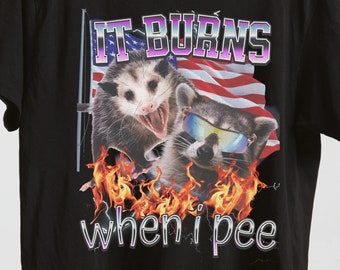 Może przedstawiać: Czarny T-shirt z grafiką przedstawiającą dwa zwierzęta, oposa i szop pracza, z płomieniami i tekstem "IT BURNS when i pee".