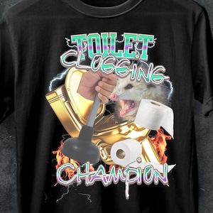 May include: A black t-shirt with a graphic of a possum holding a plunger in a golden toilet bowl. The text "Toilet Clogging Champion" is written in green and purple letters with lightning bolts.