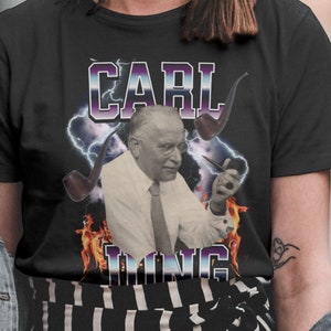 May include: Black t-shirt featuring a graphic of Carl Jung, a man in a white shirt and tie, holding a pipe. The design includes the name "CARL JUNG" in a retro font, lightning bolts, and flames.