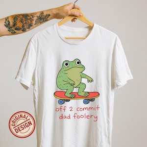Op de afbeelding: Wit T-shirt met een cartoon kikker op een skateboard. De kikker is groen en het skateboard rood. De tekst "off 2 commit dad foolery" staat in het rood. Een "ORIGINAL DESIGN" stempel is zichtbaar.