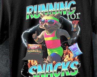 Può includere: Una maglietta nera con una grafica di un procione che indossa occhiali da sole e un completo da allenamento rosa e verde neon mentre corre con il testo "RUNNING for SNACKS" in stile retrò.