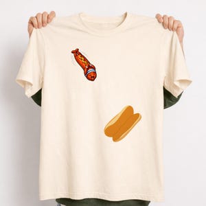以下が含まれることがあります： クリーム色のTシャツ。青いヘアバンドをした漫画のホットドッグが、2つのホットドッグバンズに向かって飛び込んでいる。ホットドッグは赤い体と黄色のマスタード。バンズは薄茶色。食べ物をテーマにした楽しいデザイン。