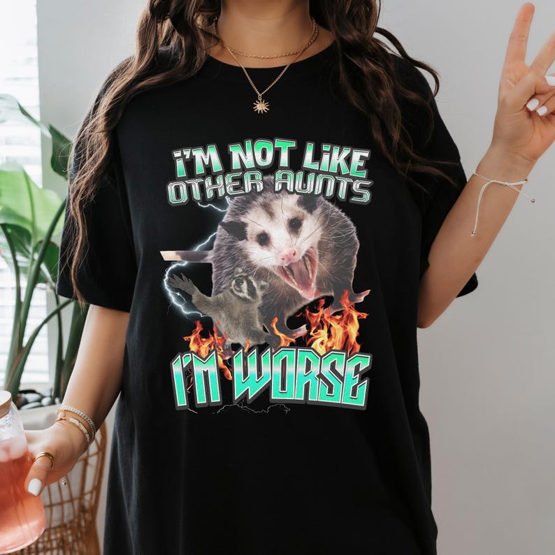 May include: A black t-shirt featuring a graphic print of a screaming opossum and a small raccoon surrounded by orange flames and blue lightning. The text reads I'M NOT LIKE OTHER AUNTS I'M WORSE in a bold, light blue, outlined font style.
