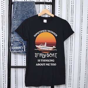 Puede incluir: Camiseta negra con un diseño de puesta de sol y un barco. El texto dice: "Sometimes I Wonder If My Boat Is Thinking About Me Too." La camiseta está colgada en una percha de madera con unos vaqueros.