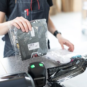 K&ouml;nnte beinhalten: Ein graues T-Shirt ist gefaltet und in einer durchsichtigen Plastikt&uuml;te verpackt. Die T&uuml;te wird von einer Hand gehalten. Das T-Shirt hat einen schwarz-wei&szlig;en grafischen Aufdruck auf der Vorderseite.