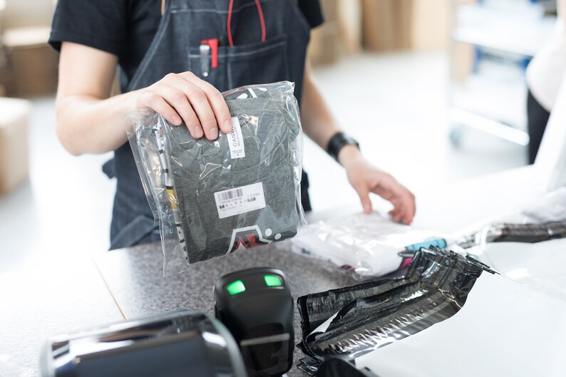 K&ouml;nnte beinhalten: Ein graues T-Shirt ist gefaltet und in einer durchsichtigen Plastikt&uuml;te verpackt. Die T&uuml;te wird von einer Hand gehalten. Das T-Shirt hat einen schwarz-wei&szlig;en grafischen Aufdruck auf der Vorderseite.