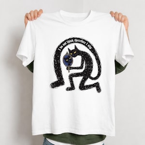 May include: White t-shirt featuring a whimsical black cat illustration. The cat, with yellow eyes, holds a mirror with a cat image. The text "I do not think therefore I nap" arches above the cat. The design is a playful, artistic statement.
