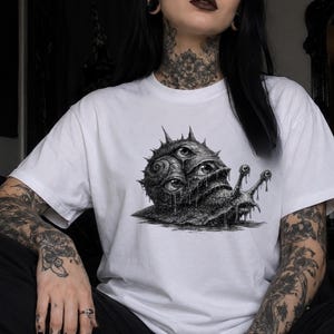 May include: A white short-sleeved t-shirt featuring a detailed black and white graphic print of a surreal snail. The snail has a shell covered in multiple realistic eyes and sharp spikes, with a dripping, textured body and two long eye stalks.