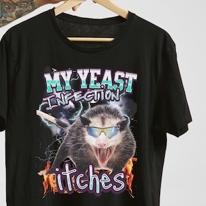 以下が含まれることがあります： サングラスをかけたオポッサムが描かれた黒いTシャツ。上部に「MY YEAST INFECTION」、下部に「itches」の文字。稲妻と炎の背景デザイン。