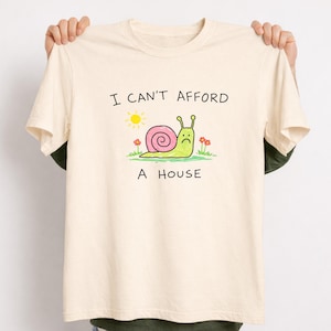 May include: Cream-colored t-shirt with the text "I CAN'T AFFORD A HOUSE". The graphic features a cartoon snail with a pink shell, a sad face, and a sun and flowers. The shirt is held up by two hands.