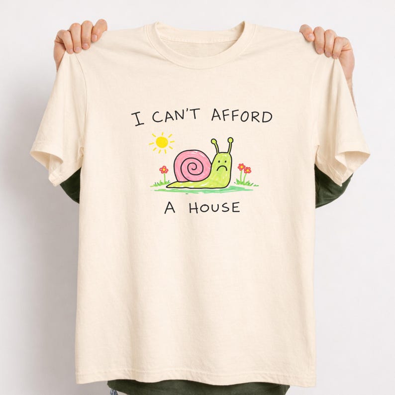 May include: Cream-colored t-shirt with the text "I CAN'T AFFORD A HOUSE". The graphic features a cartoon snail with a pink shell, a sad face, and a sun and flowers. The shirt is held up by two hands.