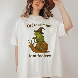 Op de afbeelding: Wit T-shirt met de tekst "Off to commit" boven een illustratie van een groene kikker met een heksenhoed die op een bruine slak rijdt. De zin "tom foolery" staat eronder.