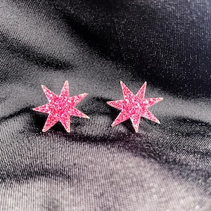 May include: Two pink glitter star-shaped earrings. Each earring has eight points and a sparkly texture. The earrings are set against a dark, textured background.