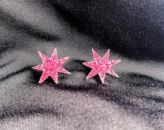 May include: Two pink glitter star-shaped earrings. Each earring has eight points and a sparkly texture. The earrings are set against a dark, textured background.