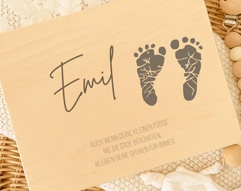 May include: A light-colored wooden plaque with the name "Emil" in cursive, and two gray baby footprints. The plaque also has text in German. The plaque rests on a white blanket and a wicker basket.