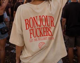 May include: A beige short-sleeved t-shirt featuring red text on the back that reads BONJOUR FUCKERS. LET THE BULLSHIT BEGIN. with a small red croissant graphic underneath the text. The shirt has a loose fit and is worn by an individual.