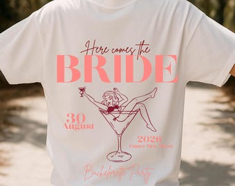May include: White t-shirt with a pink graphic design. The design includes the words "Here comes the BRIDE" and a drawing of a woman in a martini glass. The date "30 August 2026" is also included. The words "Bachelorette Party" are at the bottom.