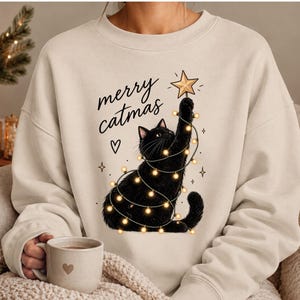 May include: A beige crewneck sweatshirt featuring a graphic of a black cat wrapped in glowing string lights, reaching up to place a gold star on top. The text "merry catmas" is printed in black script above the cat, accompanied by a small heart.