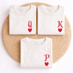 Könnte beinhalten: Drei weiße T-Shirts mit roten Spielkartensymbolen: eine Dame, ein König und eine Karte mit dem Buchstaben P, jeweils mit einem roten Herz. Die Shirts sind auf einer gewebten, kreisförmigen Unterlage gefaltet.