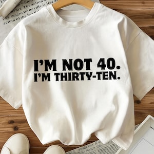Könnte beinhalten: Ein weißes T-Shirt mit dem Text "I'M NOT 40. I'M THIRTY-TEN." in fetter schwarzer Schrift. Das T-Shirt wird auf einem Holzbügel präsentiert. Das Kleidungsstück hat eine lockere Passform und scheint aus einem weichen Stoff zu sein.