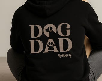 May include: Black hoodie with the words "DOG DAD" in a tan color, featuring a dog silhouette and paw print design. The name "Barney" is printed below the design in a script font. The hoodie is a casual apparel item.